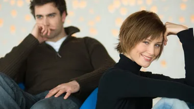 A woman smiling confidently in the foreground, with a man sitting pensively in the background.