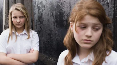 Two schoolgirls in uniform; one looks displeased with arms crossed, while the other looks downcast, standing in front.