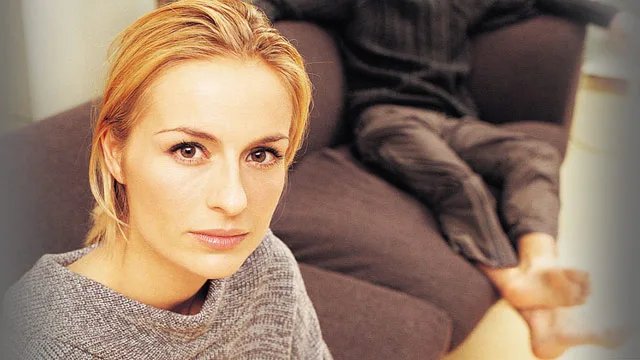 Woman with blonde hair looks intently at camera; person in background on couch, legs visible.