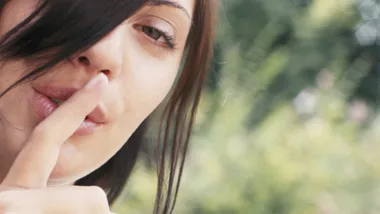 Woman with finger over lips signaling to be quiet, blurry greenery in background.