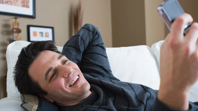 Man lounging on a couch, smiling while looking at his phone, dressed in a dark sweater with casual decor in the background.