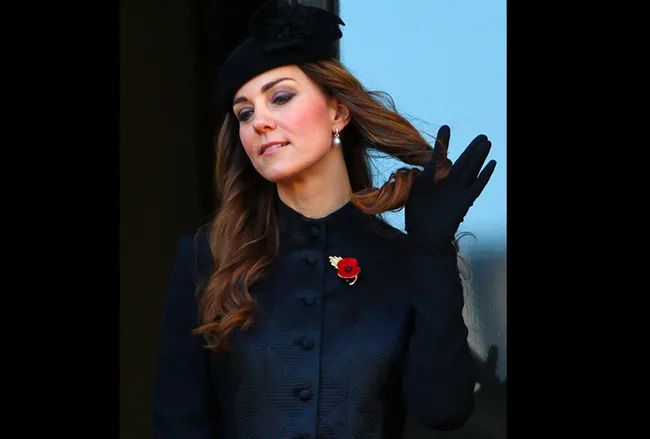 Duchess Kate Middleton in a black outfit, adjusting hair at a public event, wearing a poppy pin for Remembrance Day.