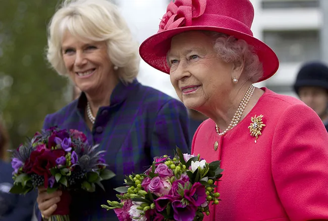 Two women smiling and holding bouquets; one in a pink hat and coat, the other in a dark jacket.