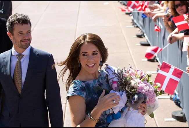 Princess Mary in a floral dress holds a bouquet, walking with a smiling man beside Danish flags in Sydney.