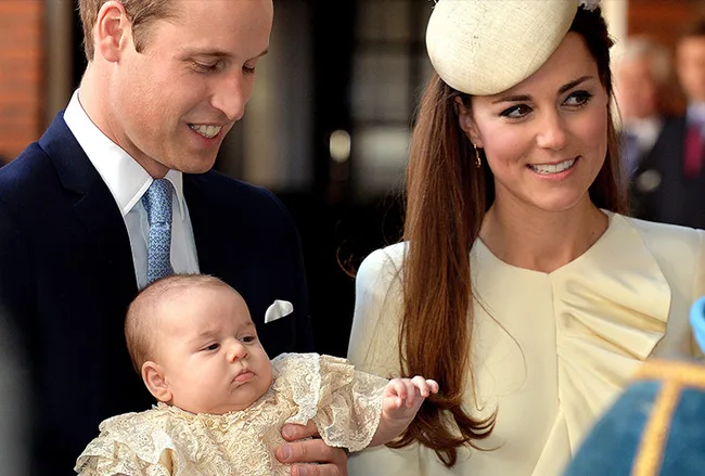 Royal couple holding their baby at a christening event, smiling and wearing elegant attire.