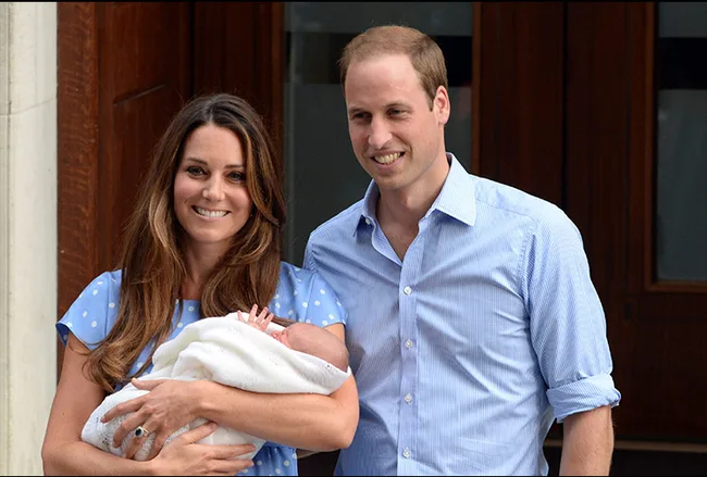 A smiling couple holding a newborn wrapped in a white blanket outside a building.