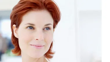 Young woman with short red hair smiling slightly, looking to her right in a bright setting.