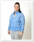A woman in a blue striped jacket and grey pants standing and smiling against a plain white background.