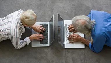 Two elderly individuals lie face-down on a carpeted floor using laptops, positioned opposite each other.