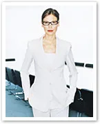 Woman in a white suit and glasses stands confidently in a modern office setting.