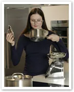 A woman in a kitchen smells inside a pot, holding the lid with several other pots visible on the counter.