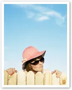Woman in pink hat and sunglasses peering over a wooden fence against a clear blue sky.