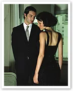 Man in a suit and woman in a black dress stand closely, engaging in a quiet conversation in a dimly lit room.
