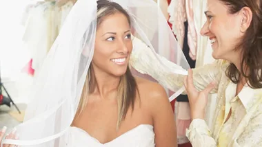 A bride smiles as another woman adjusts her veil in a bridal shop, surrounded by dresses.