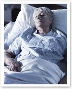 Elderly woman in hospital bed with oxygen tube, wearing a hospital gown, appears to be resting peacefully.