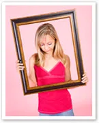 A woman in a pink top holds an empty picture frame against a pink background, looking down inside the frame.