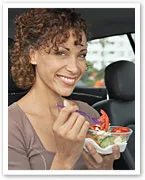 Smiling woman eating salad with a fork, sitting inside a car.