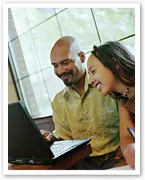 A man and woman smiling while using a laptop indoors, with a window in the background.