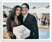 A couple posing outdoors holding a wrapped gift, with a party tent and guests in the background.