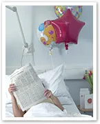 Person in hospital bed reading newspaper with Mylar balloons in background.