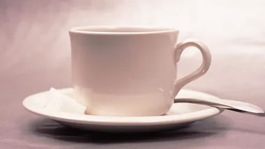 A white teacup on a saucer with a sugar cube and a spoon beside it.