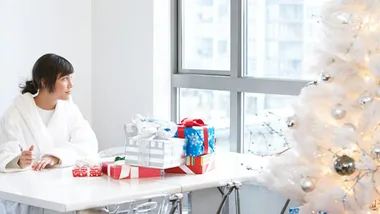 A woman in a white robe sits at a table with wrapped gifts, near a white decorated Christmas tree by a window.