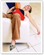 A person sleeping on a white sofa, wearing brown pants and a dark top, with a red purse and arm hanging down.