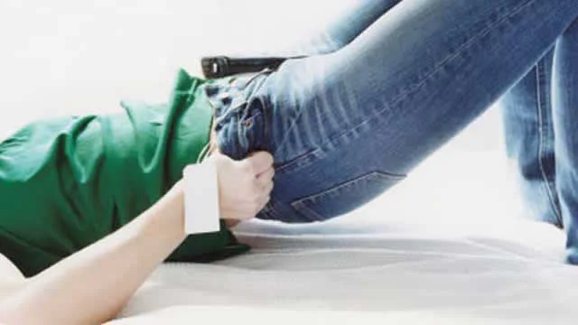 Wrist-bound person in jeans and green shirt struggles on white surface, suggesting captivity or resistance.