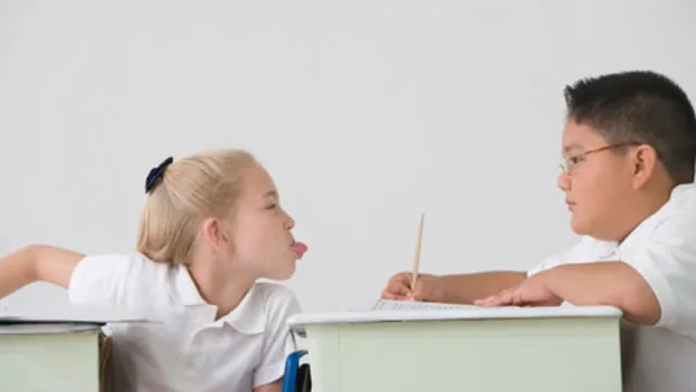 Two children in a classroom, a girl sticking her tongue out at a boy who's writing.