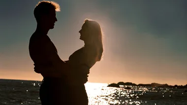 Couple silhouetted against the sunset at the beach, embracing with a serene ocean backdrop.