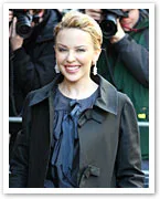 A person smiling and posing outdoors in front of photographers, wearing a black coat and earrings.