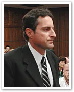 A man in a suit stands in a courtroom, with people seated in the background.