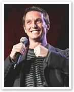 Man on stage smiling, holding a microphone in one hand, wearing a dark jacket and striped shirt.