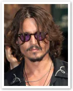 A person with shoulder-length hair, wearing sunglasses and a black shirt with light-colored stitching.