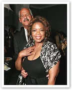 Oprah Winfrey smiling, standing with a man in a suit at a formal event.