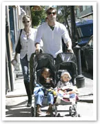 A couple walks on a street with two children in double strollers, one child holding a toy.