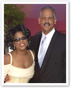 Two people smiling, one in sunglasses and formal attire, posing outdoors with trees in the background.