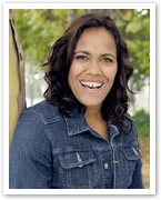 A smiling woman in a denim jacket standing outdoors with trees blurred in the background.