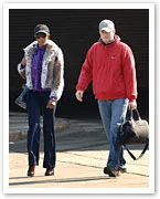 Naomi Campbell in stylish fur coat and jeans, walking with a man carrying a bag, both wearing caps.