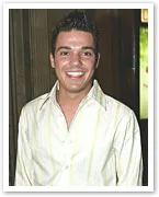 A smiling man in a light-colored striped shirt stands indoors, captured mid-torso.