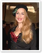 A woman in a black top and hat with long blonde hair and a bright smile, standing indoors at an event.