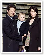 A couple with a baby pose outdoors in coats; woman holding a bag.