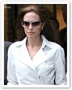 A woman in a white coat and sunglasses walking outside, partially obscured background.