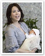 Woman smiling and holding a newborn wrapped in a blanket, standing beside greenery.