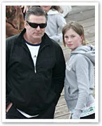 Man in black jacket and sunglasses stands with a girl in a grey hoodie on a wooden deck, with people in the background.