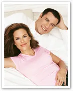 A woman in a pink shirt and a man in white shirt are lying on a bed, both smiling at the camera.