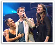 A man holding a microphone on stage with two women, one hugging him while they all face the audience under colorful stage lights.