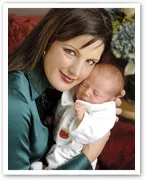 Mother in a green satin blouse smiling while holding a sleeping newborn wrapped in white.