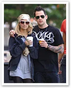A couple in casual clothing and sunglasses walking outdoors, holding drinks.