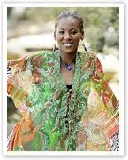 Deni Hines smiling in a colorful, patterned dress, standing outdoors with trees blurred in the background.